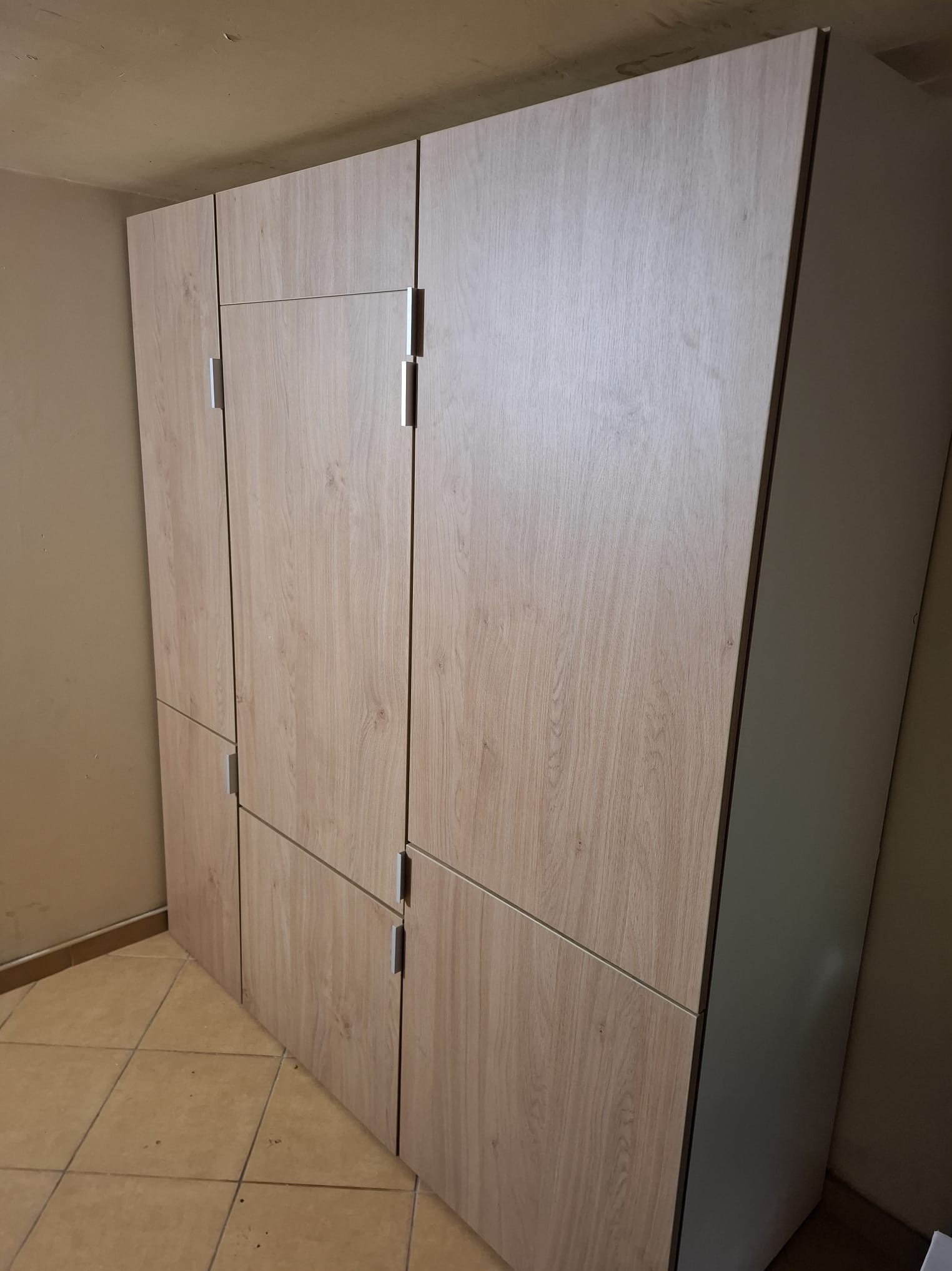 Jasna, drewnopodobna szafa z metalowymi uchwytami, podzielona na segmenty, stojąca na brązowej podłodze w pomieszczeniu o beżowych ścianach.