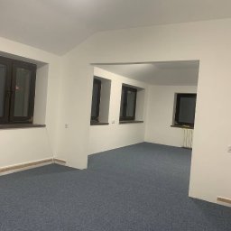 Nataliia Kosheleva Kaprys - Wnętrze po remoncie: szary dywan na podłodze, białe ściany z trzema oknami w ciemnych ramach. Widoczny fragment grzejnika. Oświetlenie sufitowe.