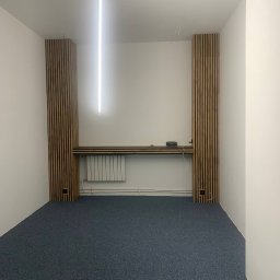 Nataliia Kosheleva Kaprys - Białe wnętrze biurowe z szarą wykładziną, drewnianymi panelami ściennymi i podłużną lampą sufitową. Widoczny grzejnik i półka. Minimalistyczny design.