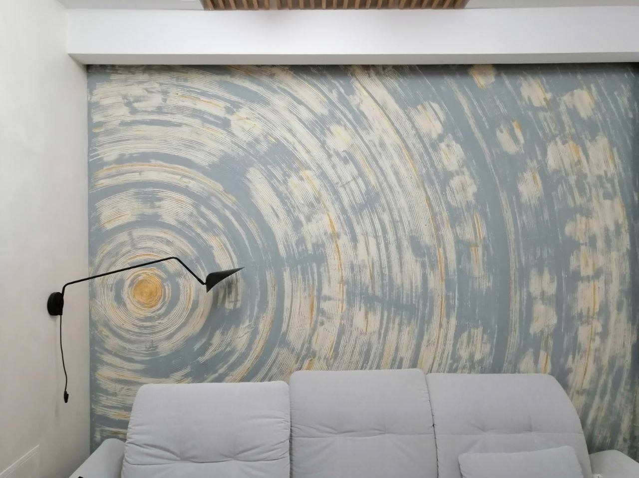 Ściana z dekoracyjnym malowaniem w odcieniach szarości i beżu, z widoczną fakturą pociągnięć pędzla, oświetlona lampą ścienną, nad szarą sofą.