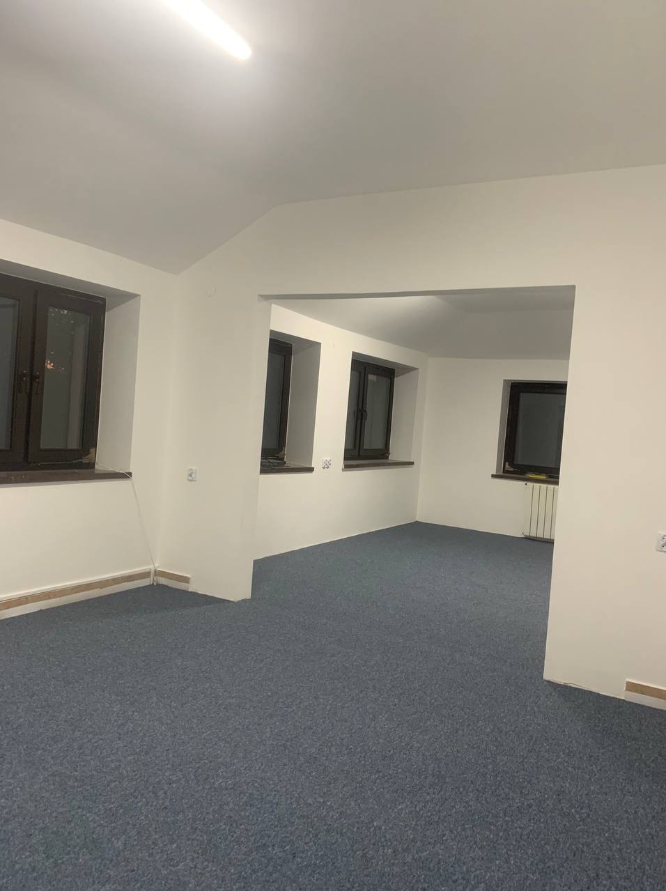 Wnętrze po remoncie: szary dywan na podłodze, białe ściany z trzema oknami w ciemnych ramach. Widoczny fragment grzejnika. Oświetlenie sufitowe.