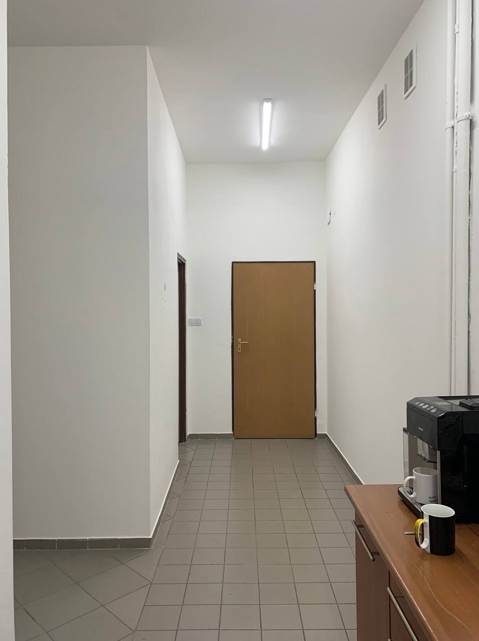Korytarz biurowy z szarą podłogą, białymi ścianami i brązowymi drzwiami. Widoczny ekspres do kawy i kubek na blacie. Oświetlenie sufitowe.