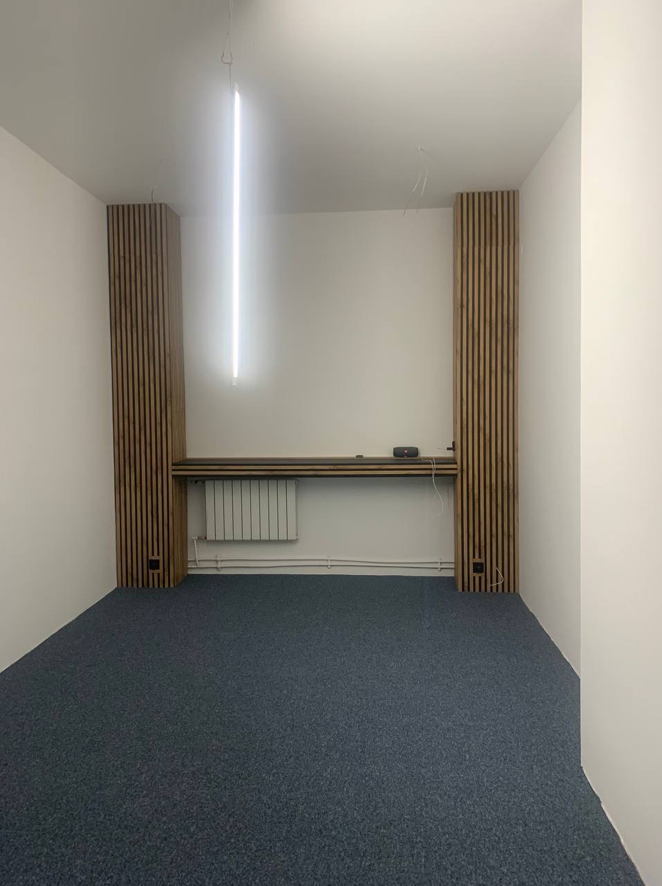 Białe wnętrze biurowe z szarą wykładziną, drewnianymi panelami ściennymi i podłużną lampą sufitową. Widoczny grzejnik i półka. Minimalistyczny design.