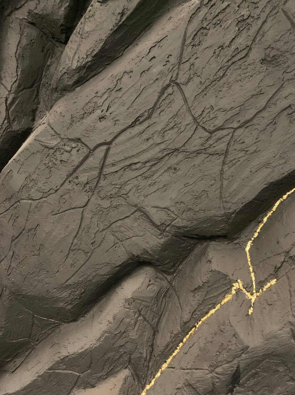 Szara, teksturowana ściana z efektem kamienia, podkreślona złotymi żyłkami, imitująca naturalne pęknięcia. Dekoracyjny element wykończenia wnętrza.