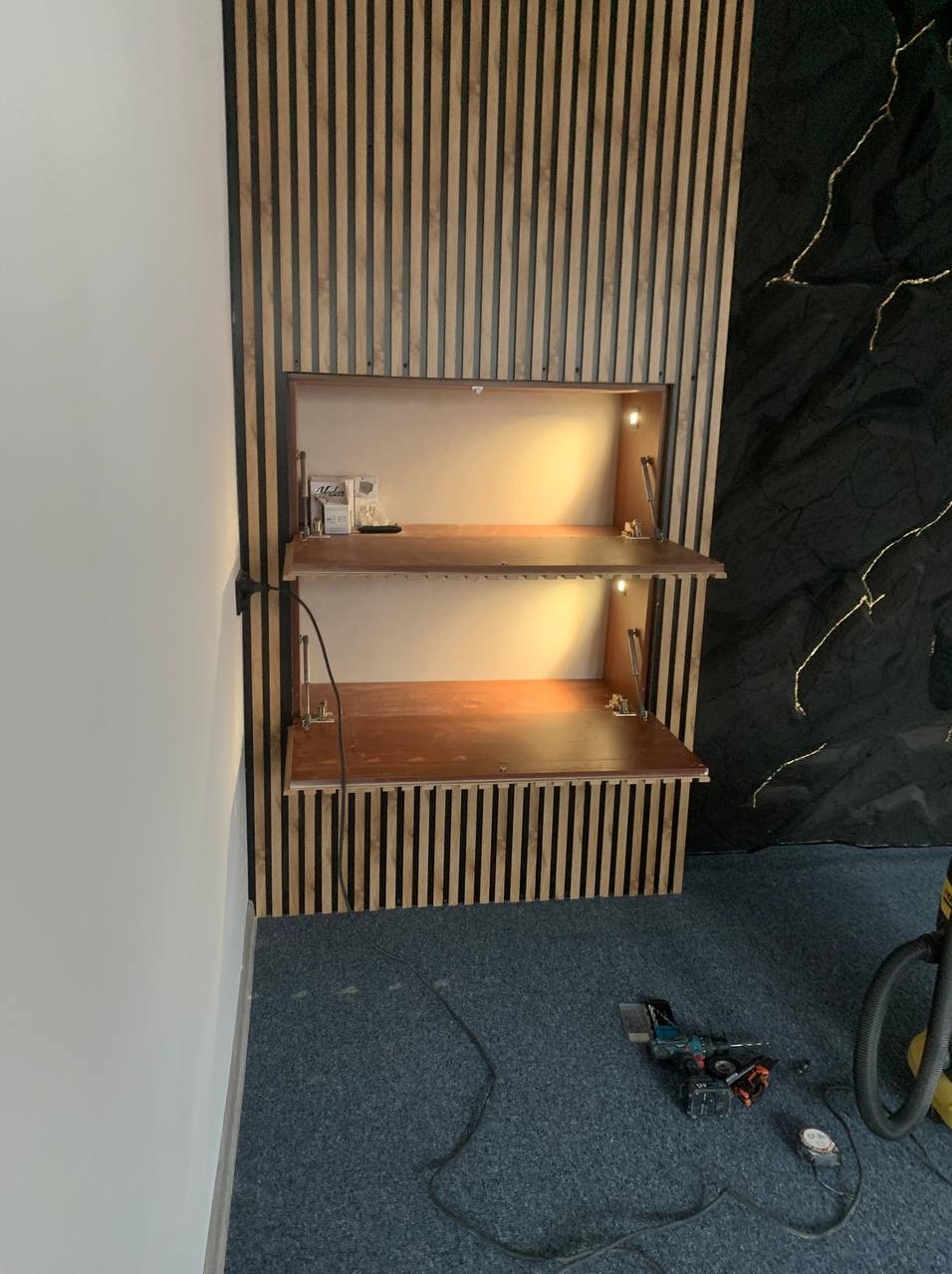 Stylowa zabudowa ścienna z drewnianymi lamelami i otwieranymi półkami z oświetleniem LED, Częstochowa. Widoczne narzędzia na podłodze.