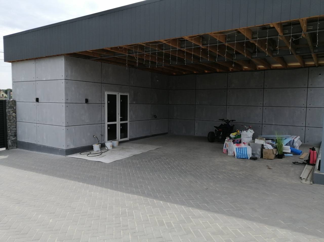 Wykończona wiata garażowa z szarymi panelami elewacyjnymi i drewnianą konstrukcją dachu. Widoczne białe drzwi, kostka brukowa i akcesoria budowlane.