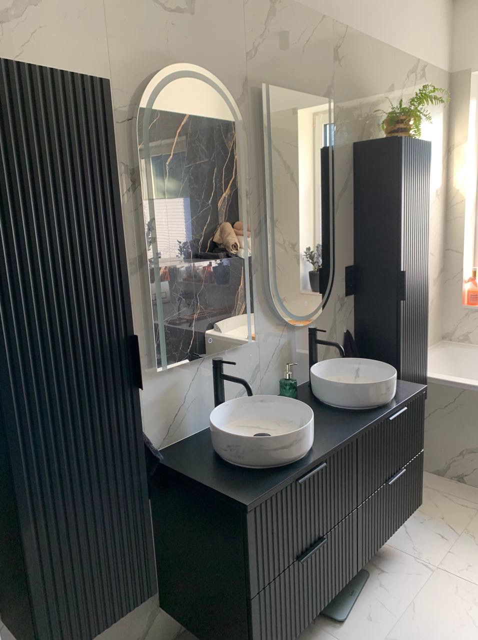 Nowoczesna łazienka z czarną szafką pod umywalki, marmurowymi umywalkami i podświetlanymi lustrami owalnymi. Minimalistyczny design z rośliną w tle.