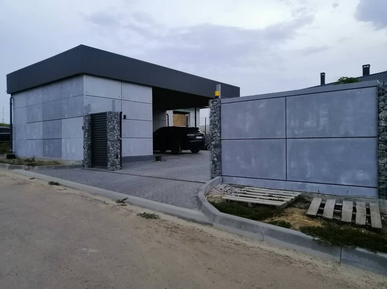 Nowoczesna brama wjazdowa z szarego betonu architektonicznego i gabionów, prowadząca do garażu z czarnym samochodem w tle, kostka brukowa na podjeździe.