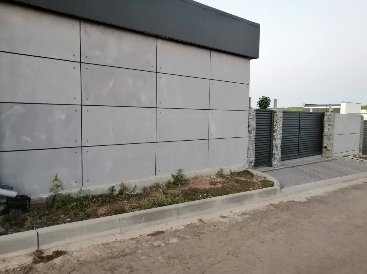 Elewacja budynku z szarych paneli i brama wjazdowa z gabionowymi słupkami. Minimalistyczny design, nowoczesne wykończenie zewnętrzne.