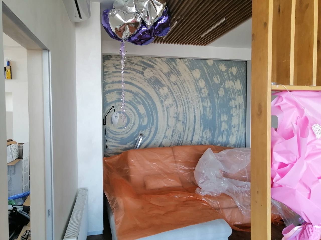 Pokój przed remontem: sofa zabezpieczona folią, dekoracyjna ściana z motywem okręgów, balon w kształcie serca pod sufitem. Widoczne przygotowania do prac remontowych.