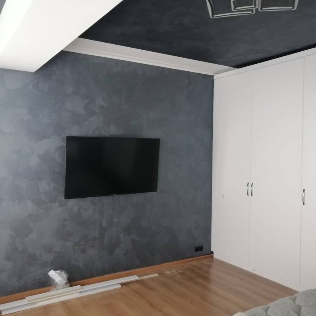 Ściana z efektem dekoracyjnym w odcieniach szarości, na niej zawieszony telewizor. Obok biała szafa wnękowa. Na podłodze listwy przypodłogowe.