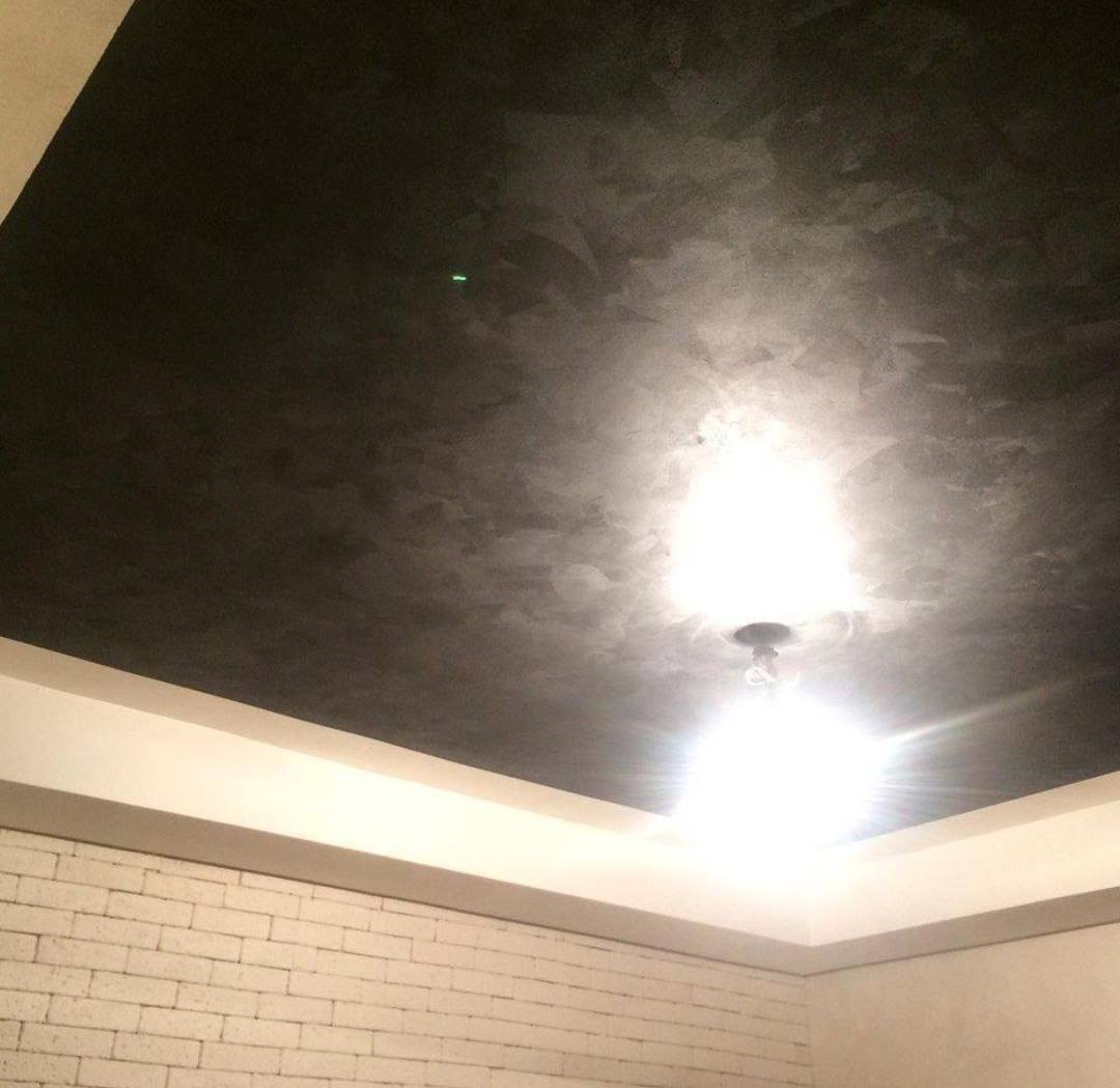 Ciemne, dekoracyjne wykończenie sufitu z widoczną lampą, białe listwy przysufitowe i fragment ściany z cegły w dolnej części kadru. Nietypowe oświetlenie.