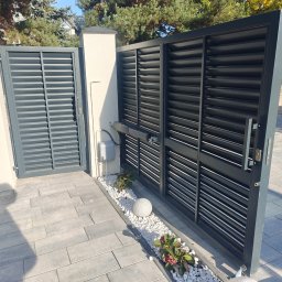 Dron Elektro - Nowoczesna, grafitowa brama lamelowa z automatyką, otwierana na posesję wyłożoną szarą kostką brukową. Widoczny element dekoracyjny w postaci białych kamieni i roślin.