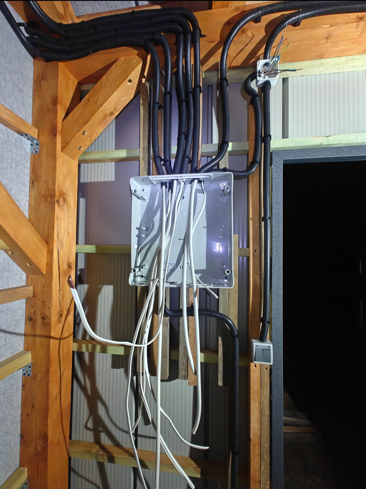 Rozdzielnica elektryczna z przewodami w peszlach, zamontowana na drewnianej konstrukcji ściany. Widoczne kable i puszka instalacyjna. Etap prac instalacyjnych.