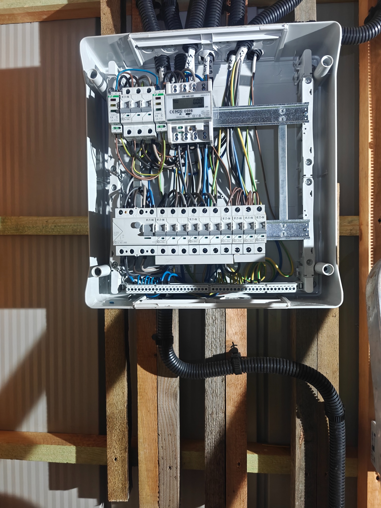 Otwarta, biała skrzynka rozdzielcza z widocznymi bezpiecznikami, przewodami i licznikiem, zamontowana na drewnianej konstrukcji ściany. Widoczne kable w peszlach.