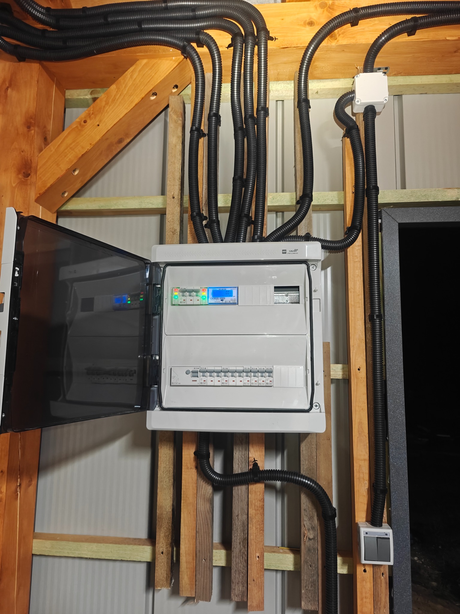 Rozdzielnica elektryczna z otwartymi drzwiczkami, widoczne kable w peszlach, zamontowana na drewnianej konstrukcji szkieletowej budynku. Widoczny włącznik światła.