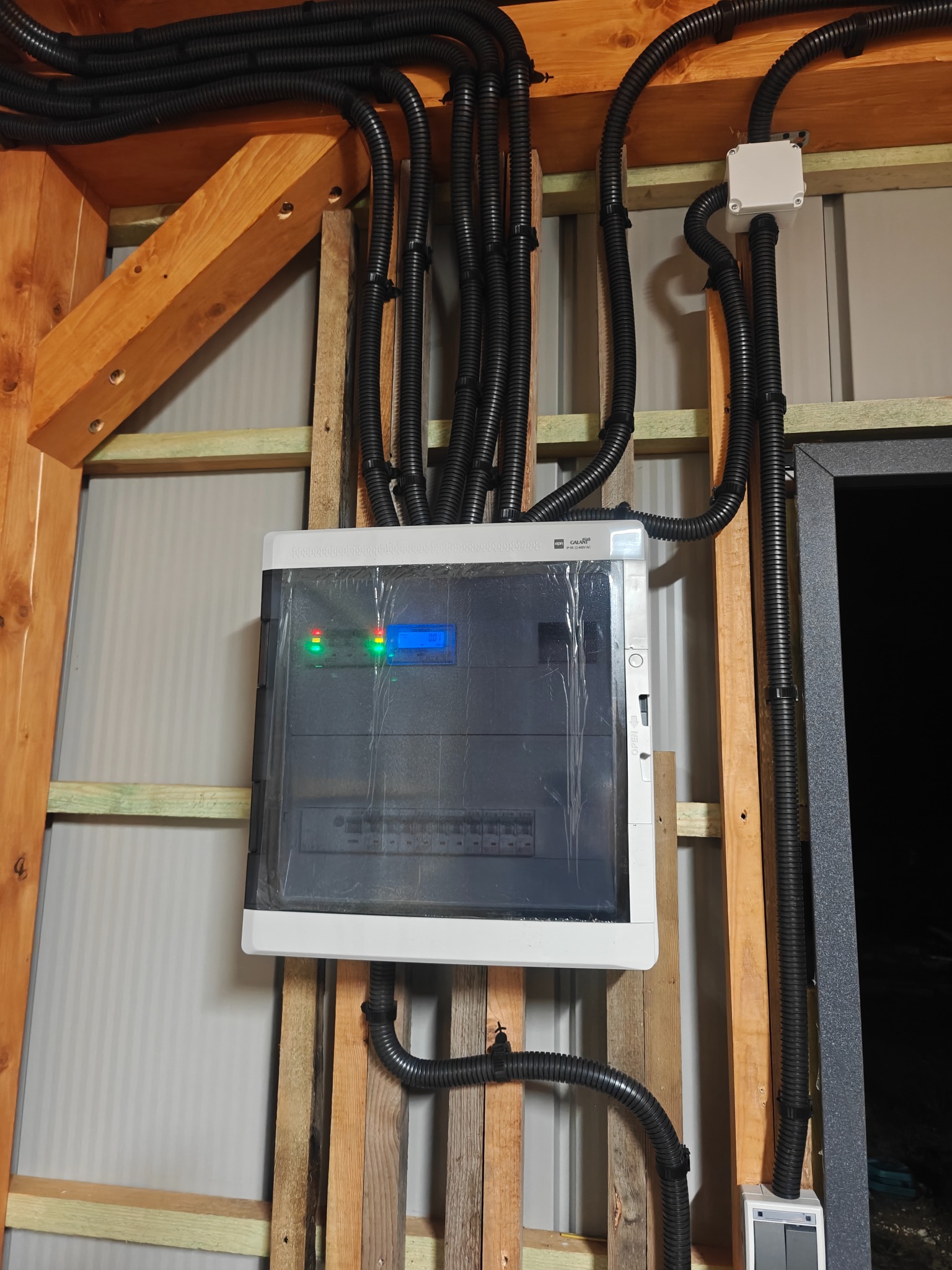 Rozdzielnica elektryczna z podłączonymi przewodami w peszlach, zamontowana na drewnianej konstrukcji. Widoczne diody sygnalizacyjne i wyświetlacz.