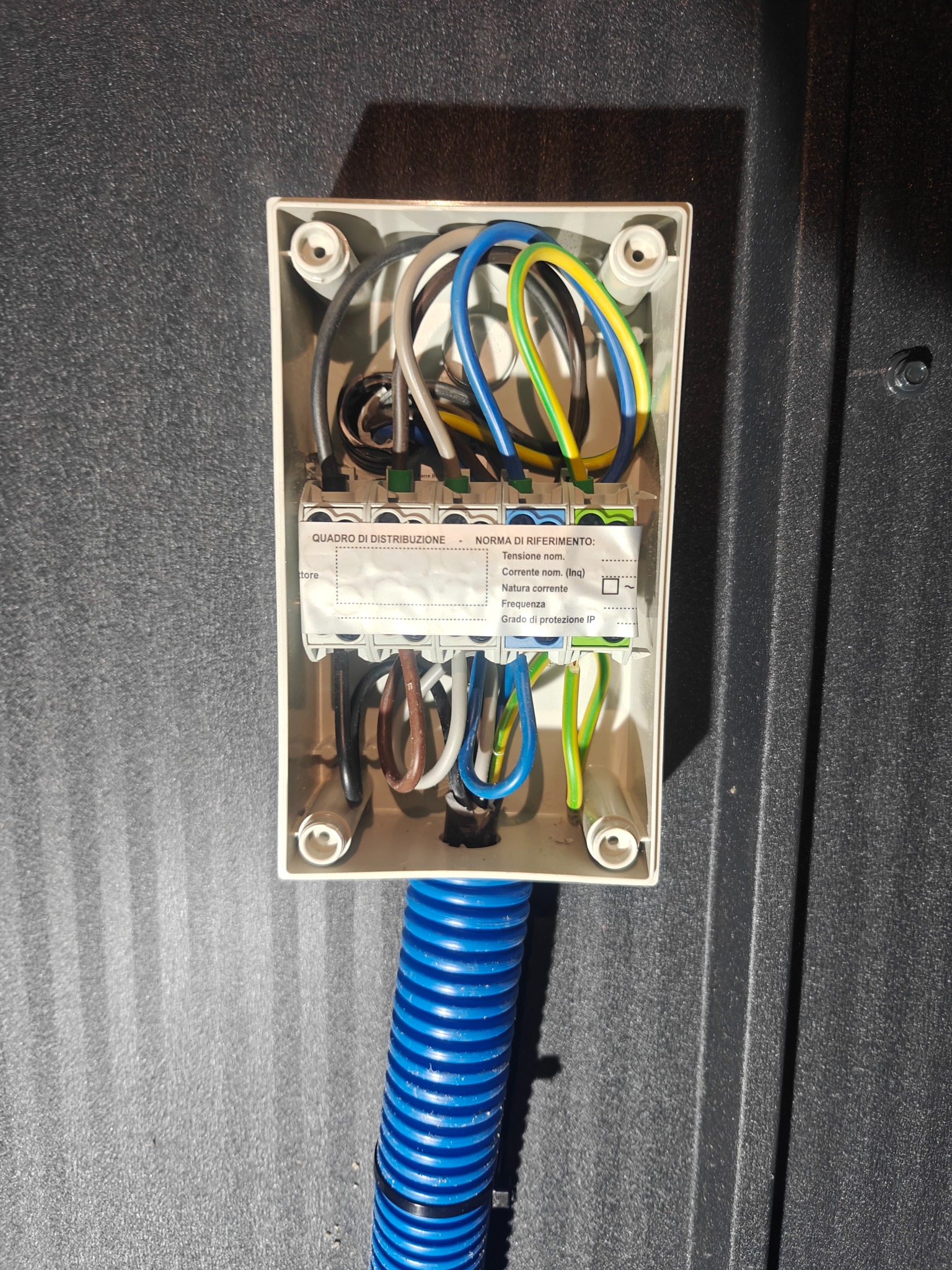 Otwarta puszka elektryczna z widocznymi przewodami w różnych kolorach, podłączona do niebieskiej rury karbowanej na tle ciemnej, teksturowanej powierzchni.