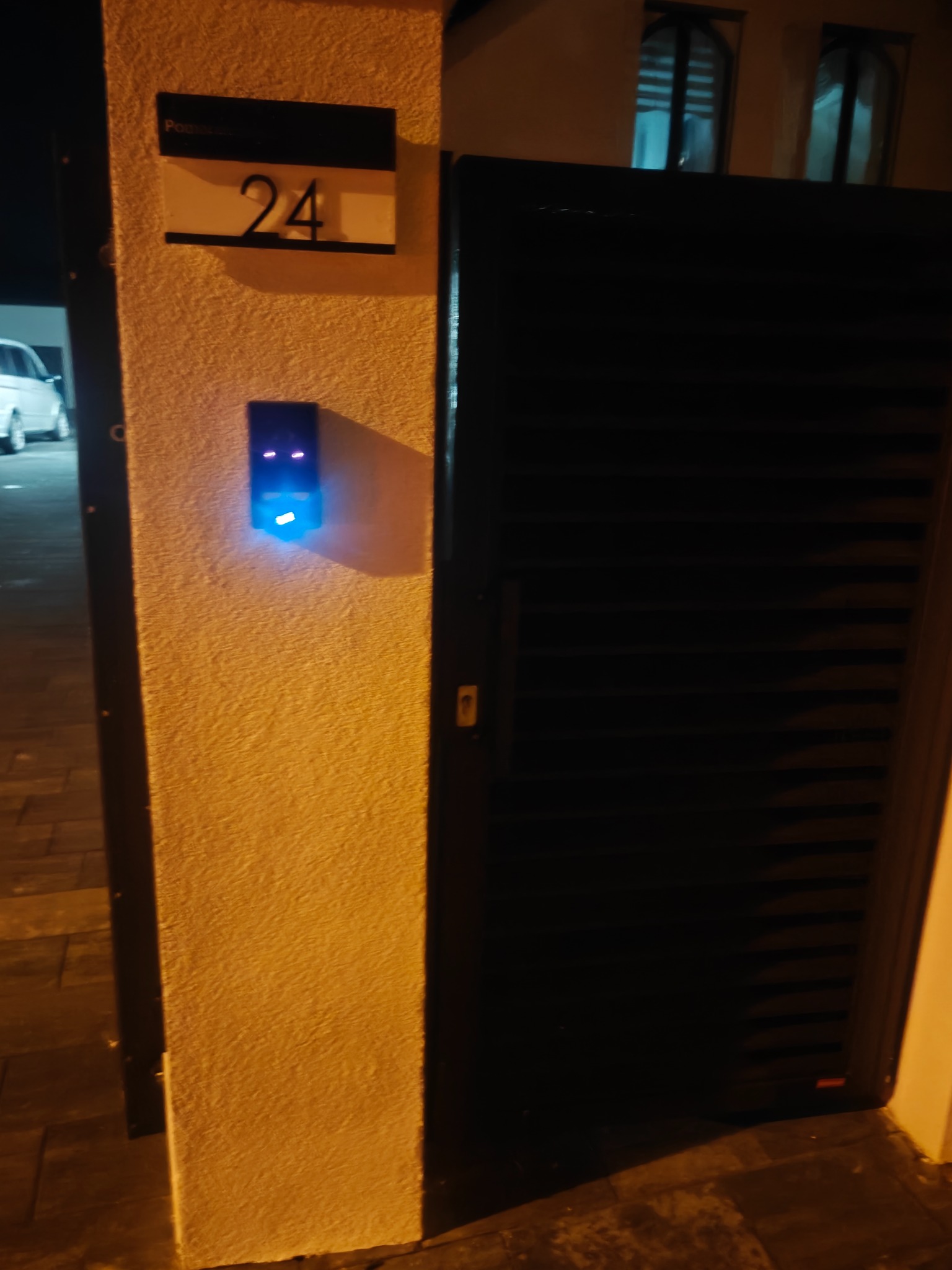 Brama posesji z numerem 24 i zamontowanym, podświetlonym na niebiesko domofonem. Widoczne okna w tle. Ujęcie nocne.