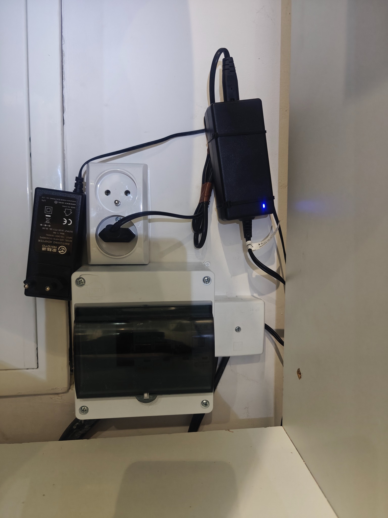 Rozdzielnica elektryczna z gniazdkiem i zasilaczami na ścianie. Widoczne przewody i elementy instalacji. Białe tło ściany.