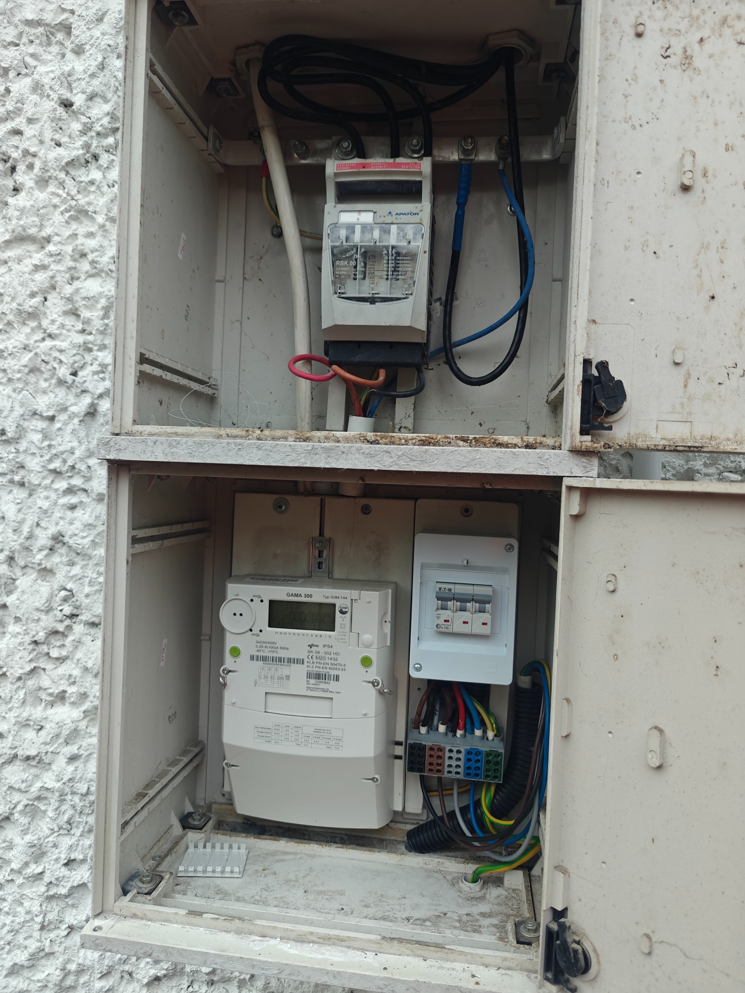 Skrzynka z licznikiem elektrycznym i zabezpieczeniami, widoczne przewody i elementy instalacji. Biała, chropowata ściana w tle.