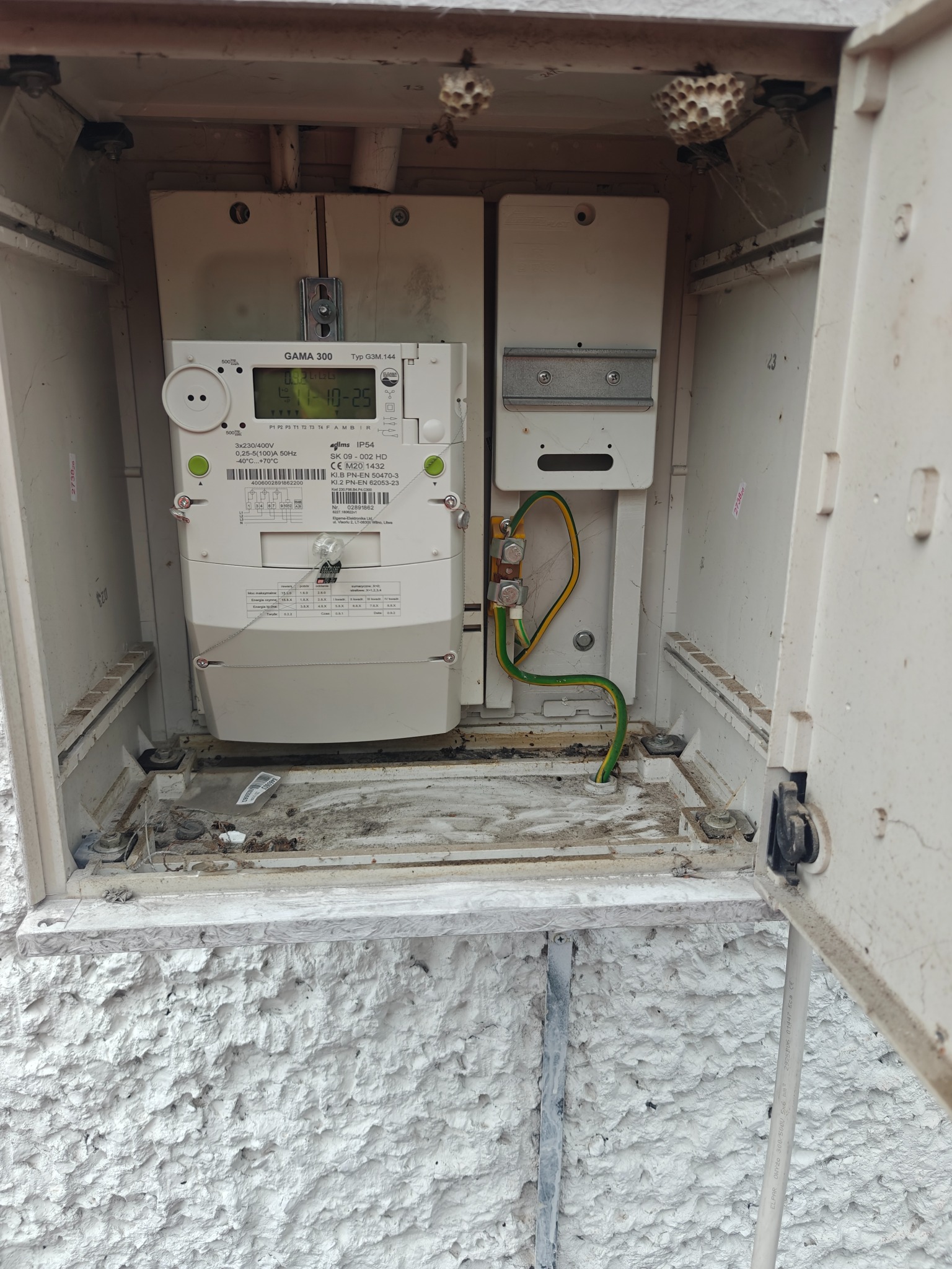 Skrzynka z licznikiem elektrycznym GAMA 300, widoczne przewody uziemiające. Na górze gniazda os. Biała, chropowata ściana zewnętrzna.