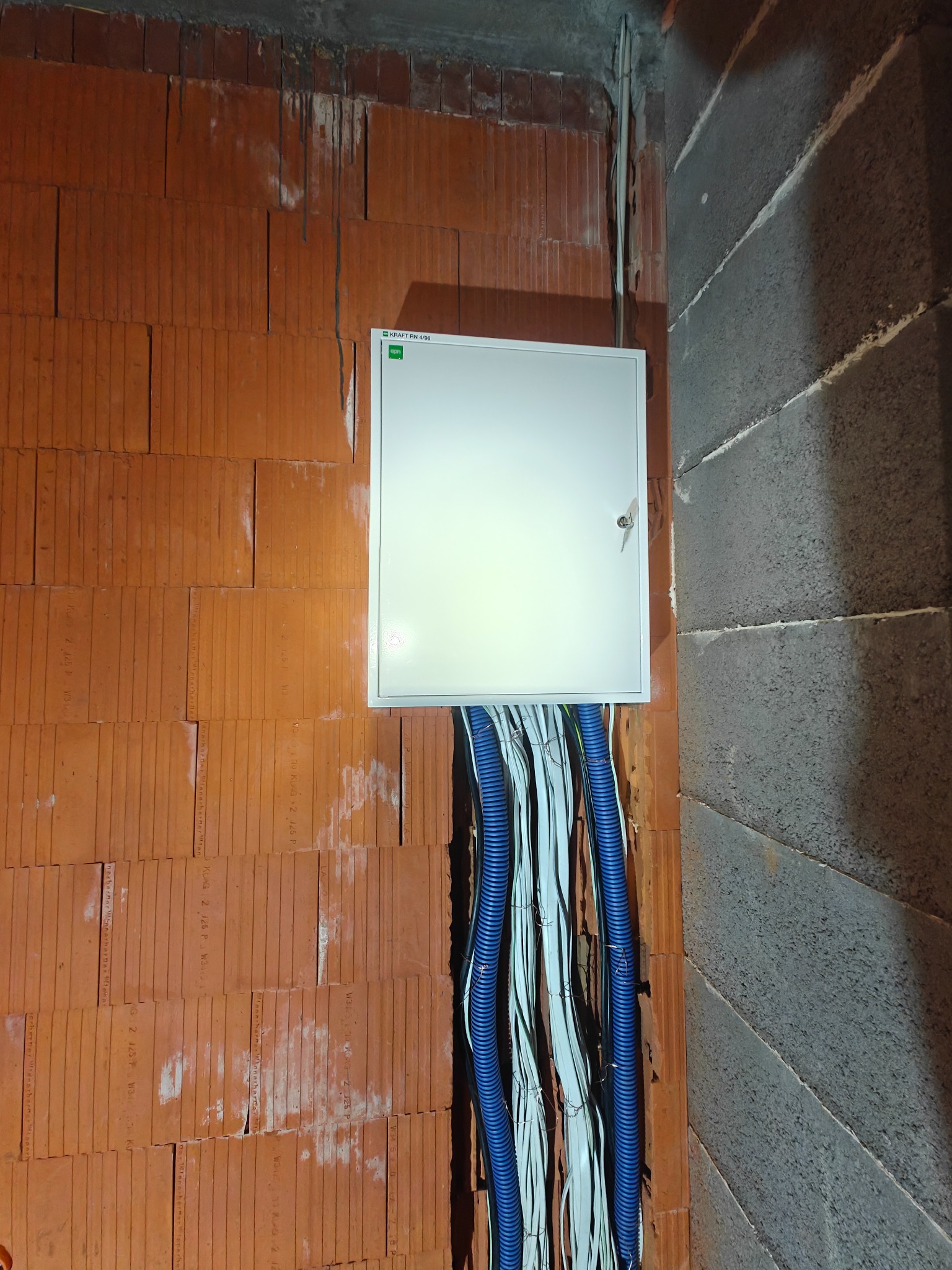 Biała skrzynka elektryczna zamontowana na ceglanej ścianie z widocznymi niebieskimi i białymi kablami w peszlach. Surowy stan wykończenia wnętrza.