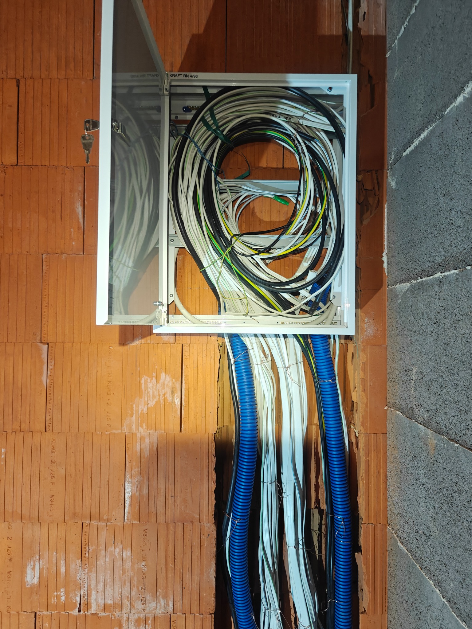 Otwarta skrzynka z kablami w ścianie z cegły i betonu. Widoczne kable w kolorach białym, czarnym, zielonym i niebieskim, wychodzące z rurek. Kluczyk w zamku skrzynki.
