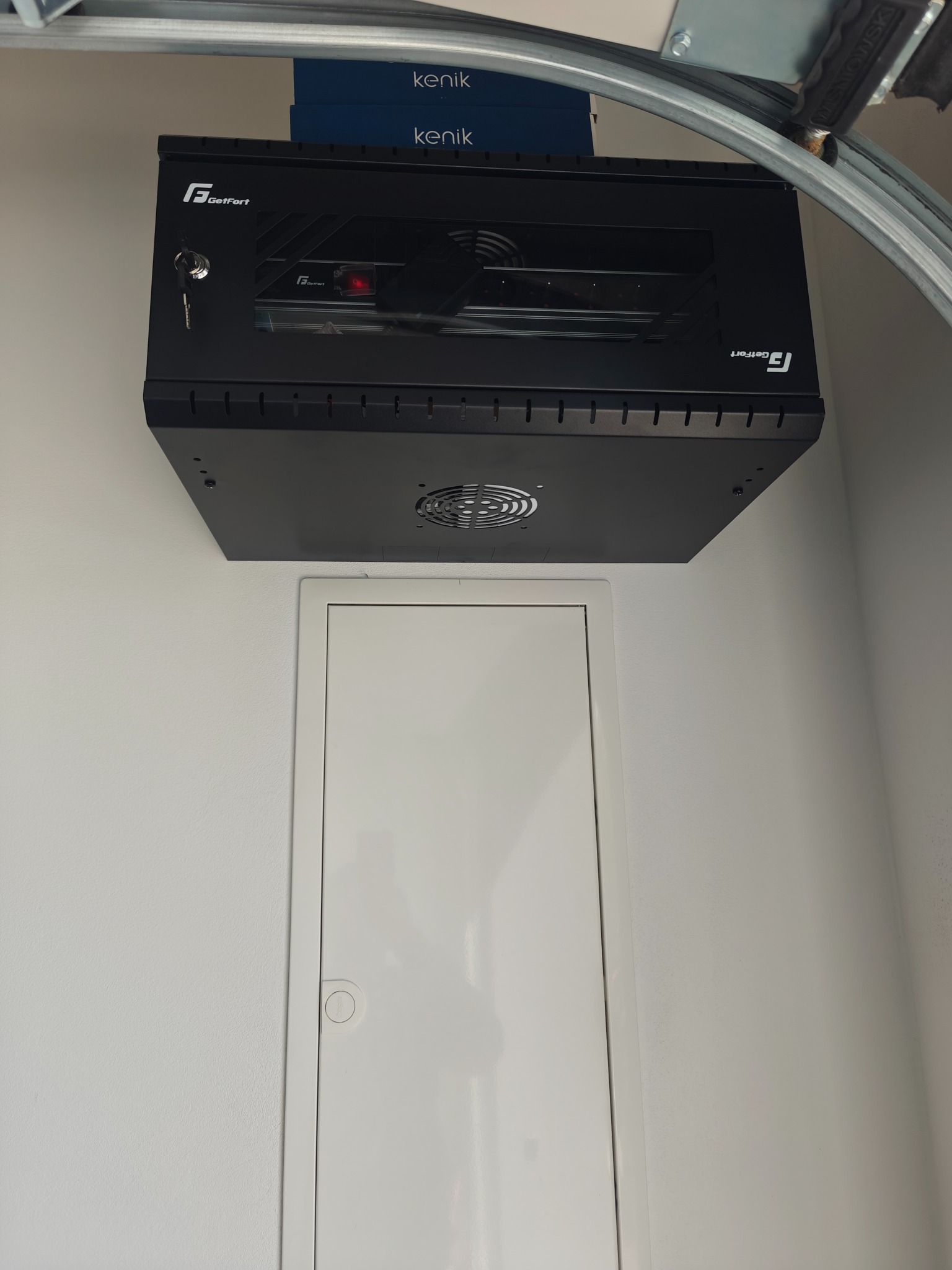 Czarna szafa rack GetFort zamontowana pod sufitem garażu, widoczna obok biała klapa rewizyjna i fragment prowadnicy bramy garażowej.