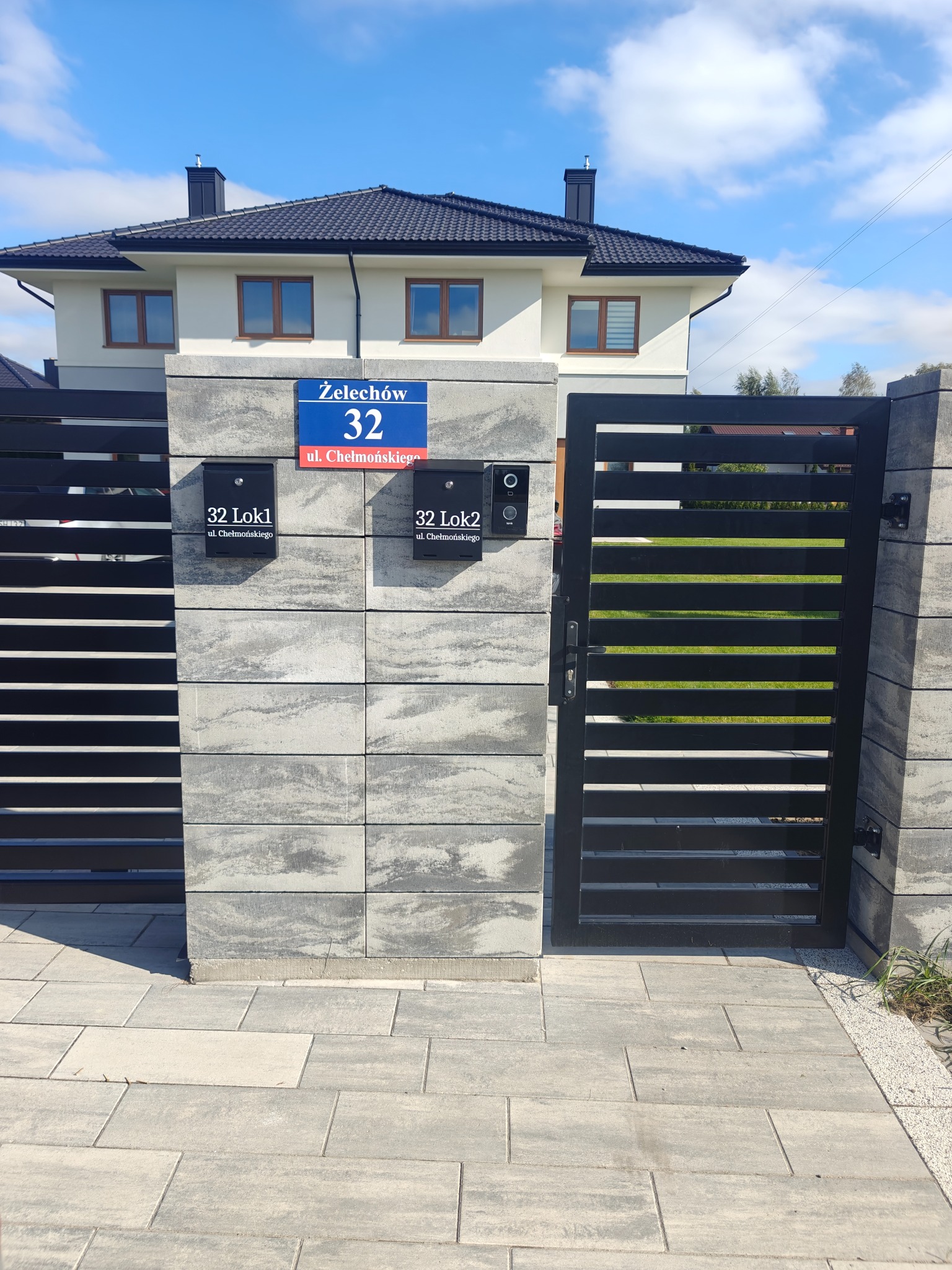 Nowoczesna brama z poziomymi szczeblami i szarego betonu słupkami, z domofonami i skrzynkami pocztowymi, na tle domu z ciemnym dachem i błękitnego nieba.