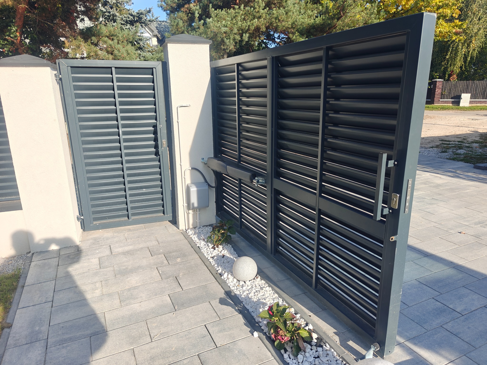 Nowoczesna, grafitowa brama lamelowa z automatyką, otwierana na posesję wyłożoną szarą kostką brukową. Widoczny element dekoracyjny w postaci białych kamieni i roślin.