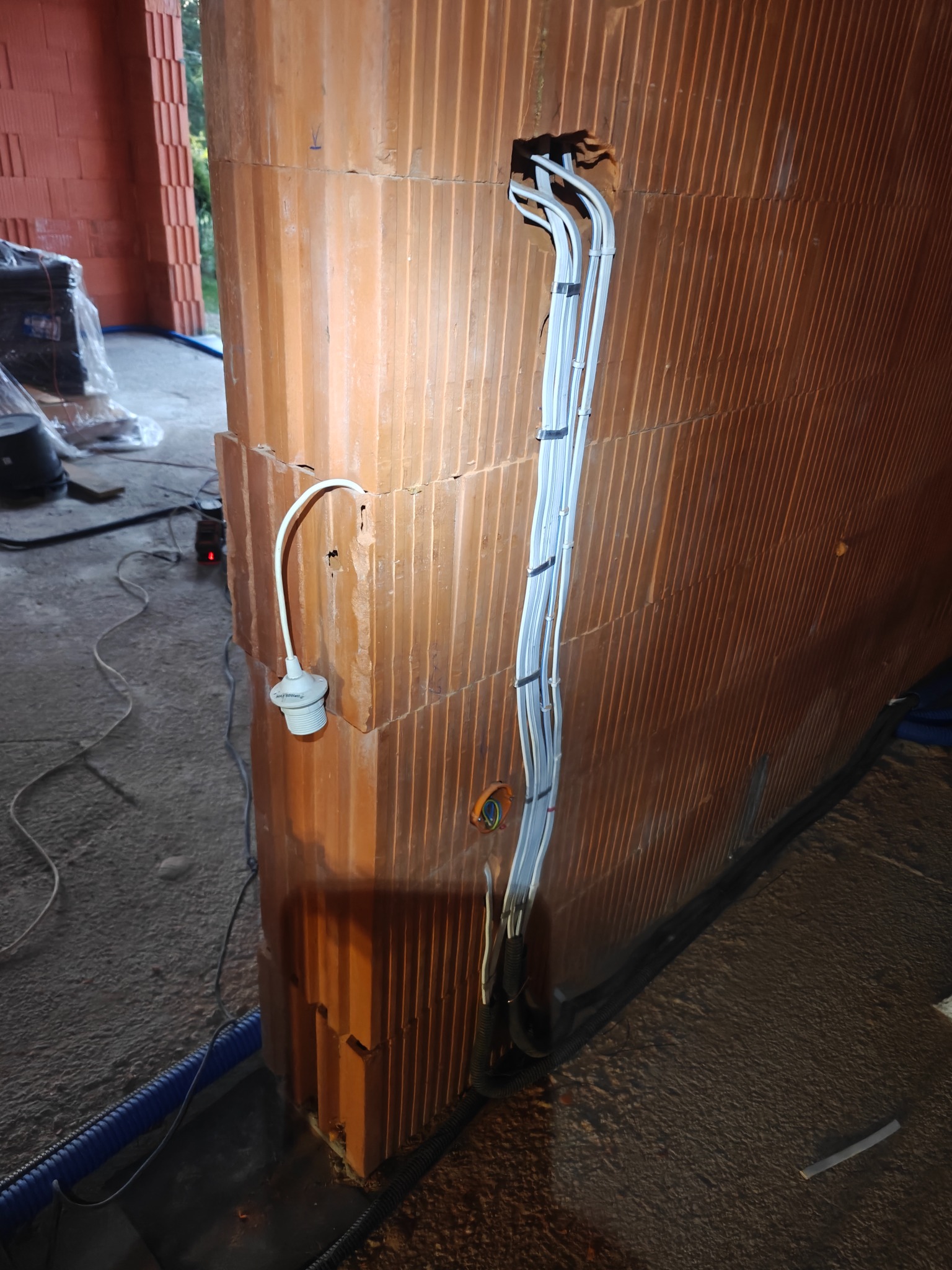 Instalacja elektryczna w ścianie z pustaków ceramicznych. Widoczne kable prowadzone w bruzdach i tymczasowe oświetlenie z oprawką. Prace w trakcie budowy.