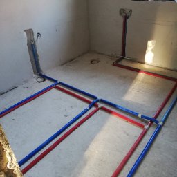Mat-San Mateusz Grenda - Rozłożone rury instalacji wodnej (czerwone i niebieskie) na surowej posadzce w pomieszczeniu przed wylaniem wylewki. Widoczne podejścia do instalacji na ścianach.
