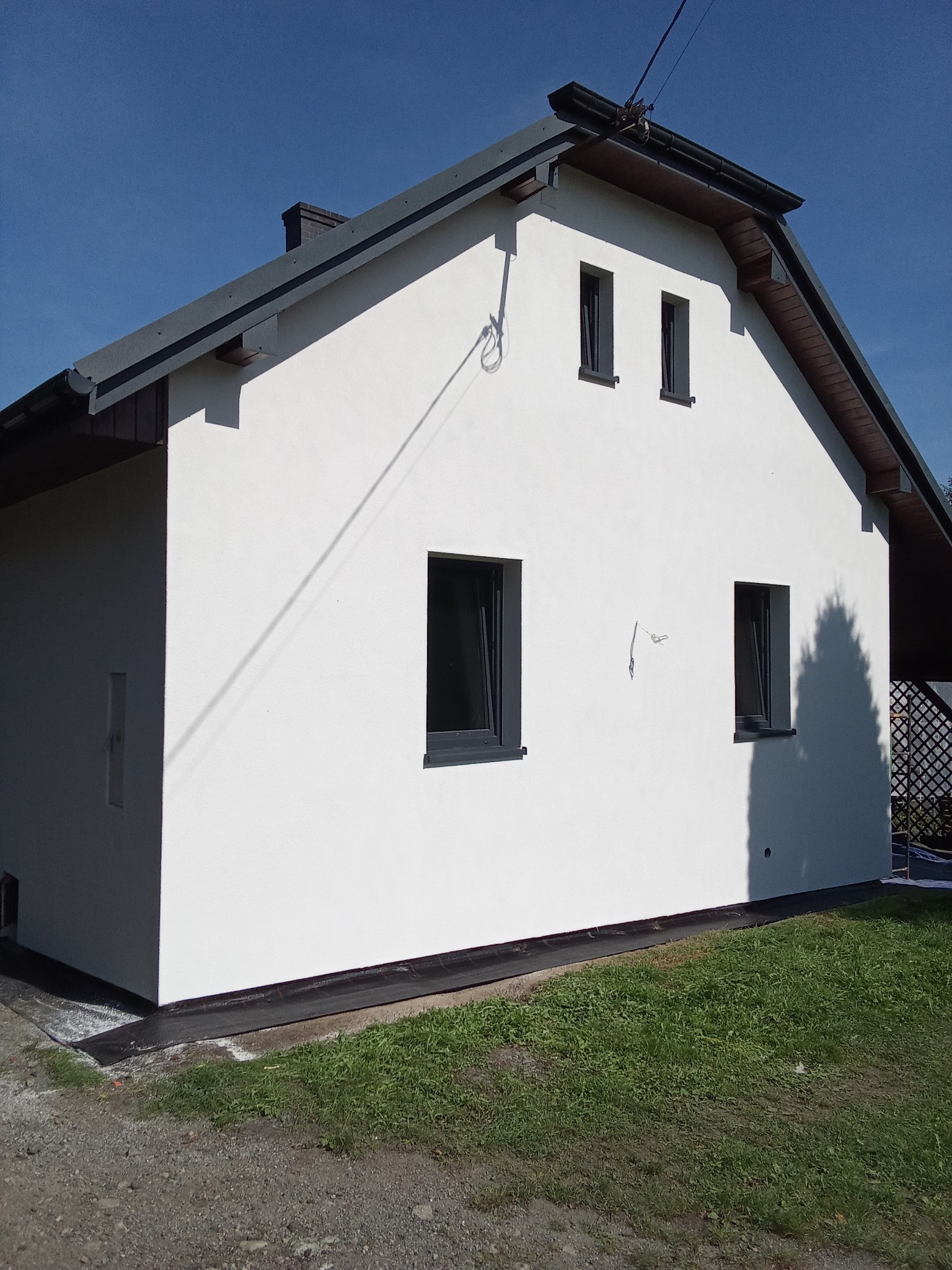 Biała elewacja domu jednorodzinnego z ciemnymi oknami i dachem, widok z boku na tle błękitnego nieba i zielonego trawnika. Minimalistyczny styl.