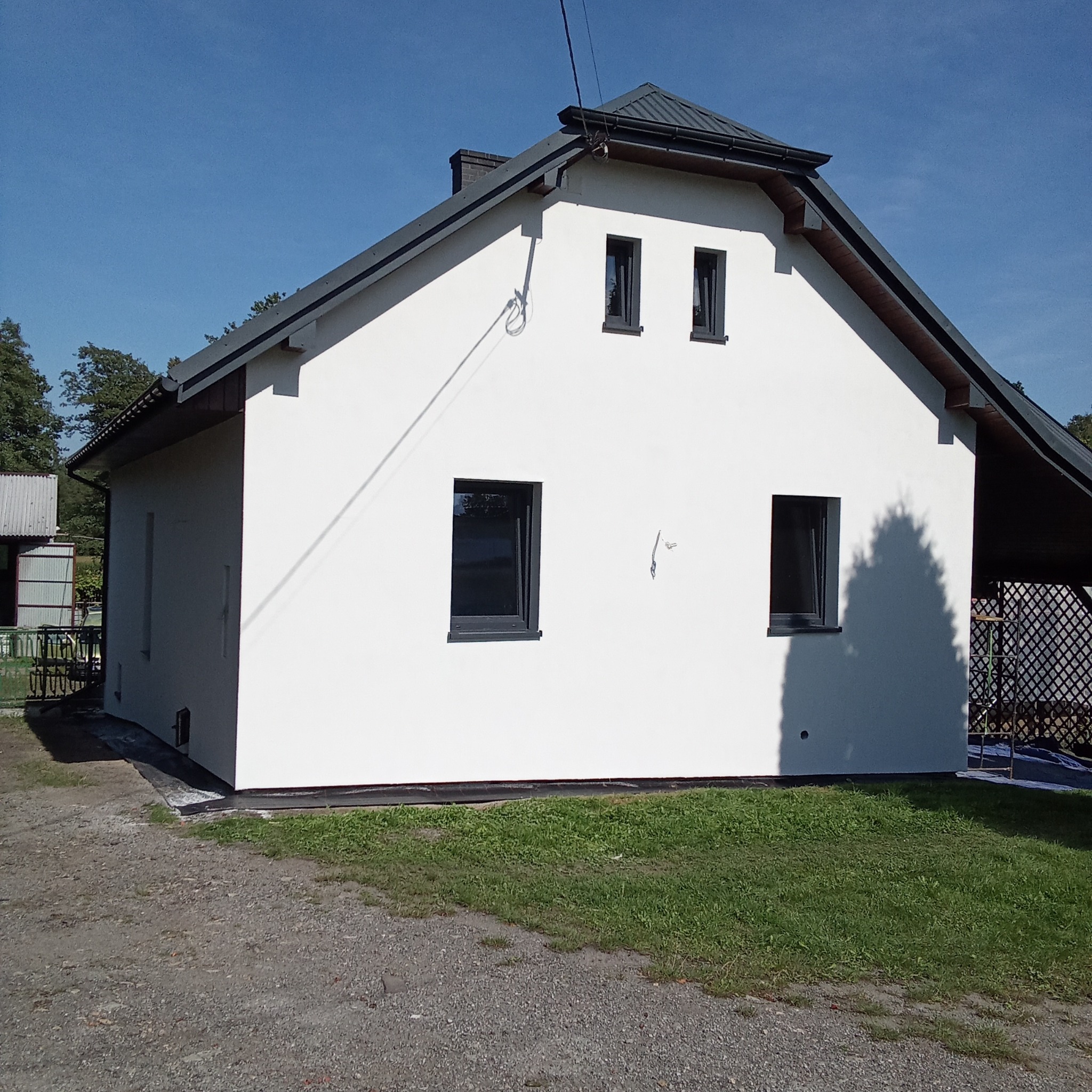 Biały dom jednorodzinny z ciemnymi oknami i dachem, świeżo po malowaniu elewacji, widok z zewnątrz w słoneczny dzień. Widoczne detale wykończenia.