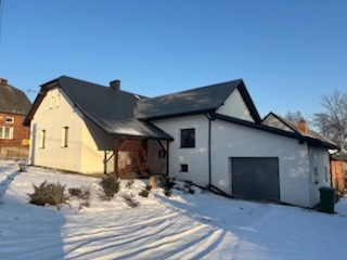 Biały dom z szarym dachem w zimowej scenerii, widok z dołu, boczny garaż, drewniana weranda, śnieg na trawniku, błękitne niebo.