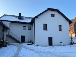 Biały, dwukondygnacyjny dom z ciemnymi oknami i drzwiami, pokryty śniegiem, widziany z perspektywy lekko z dołu, w słoneczny dzień.