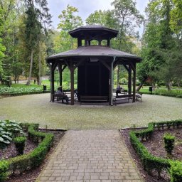 SELEN S.C. KAZIMIERZ ŁAPCZUK, MAREK MATUSIK PRODUKCJA TĘŻNI SOLANKOWYCH I GROT SOLNYCH - Ośmioboczna altana z ciemnego drewna w parku, z osobami siedzącymi na ławkach wewnątrz. Brukowana ścieżka prowadzi do altany, otoczonej zielenią i drzewami.