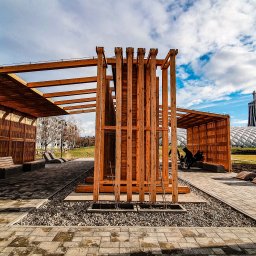 SELEN S.C. KAZIMIERZ ŁAPCZUK, MAREK MATUSIK PRODUKCJA TĘŻNI SOLANKOWYCH I GROT SOLNYCH - Nowoczesna, drewniana konstrukcja z zadaszeniem w parku miejskim. Elementy z drewna tworzą ażurową ścianę. W tle widoczny charakterystyczny budynek.