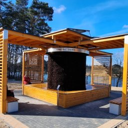 SELEN S.C. KAZIMIERZ ŁAPCZUK, MAREK MATUSIK PRODUKCJA TĘŻNI SOLANKOWYCH I GROT SOLNYCH - Drewniana konstrukcja solankowa z napisem 'Chwytaj Dzień' w parku, z widocznymi elementami drewnianymi i osobami siedzącymi na ławkach. Jasne, słoneczne oświetlenie.