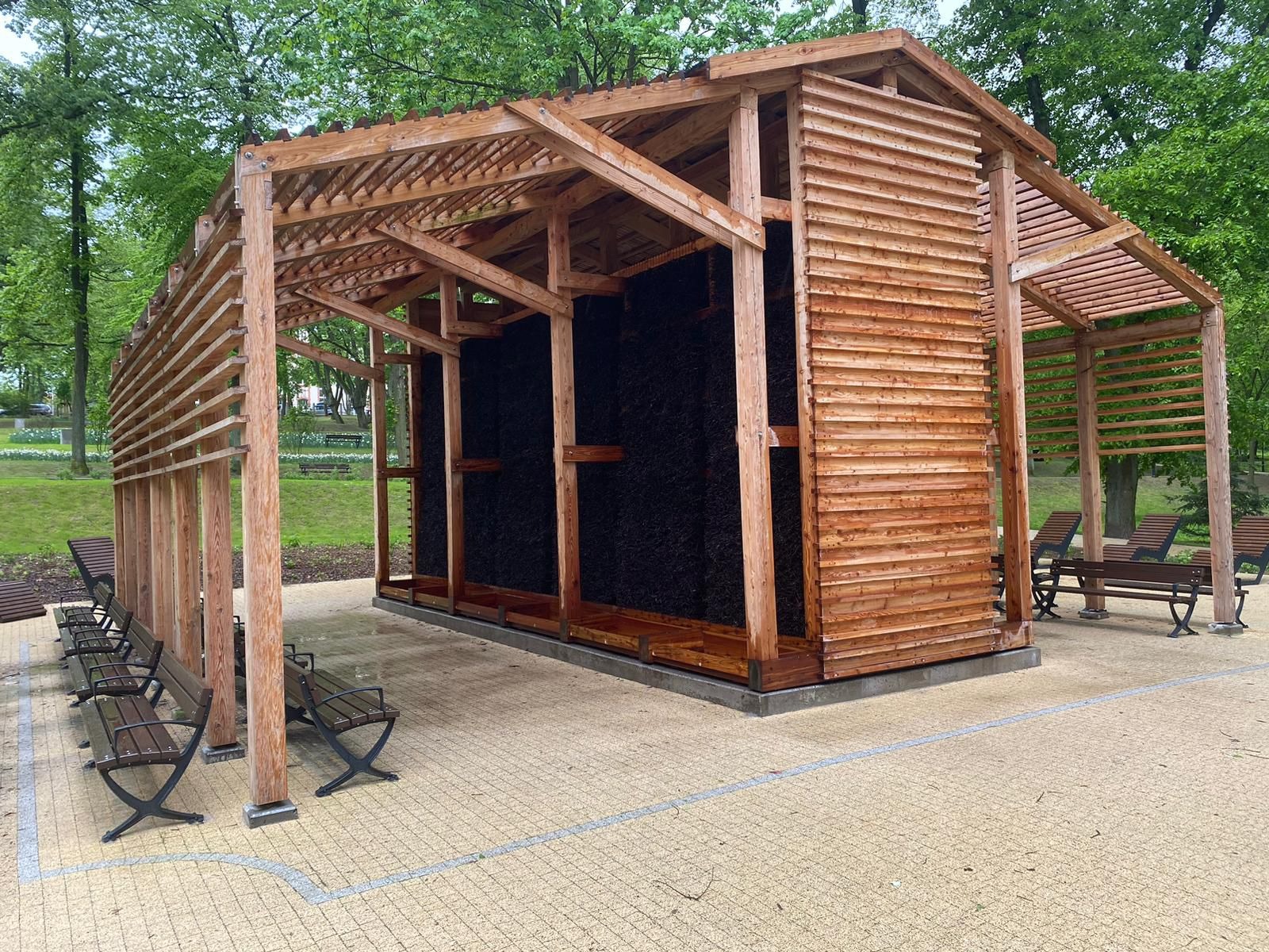 Drewniana tężnia solankowa z ażurowym dachem w parku, obok ławki. Konstrukcja z jasnego drewna, wypełniona ciemnym materiałem filtracyjnym.