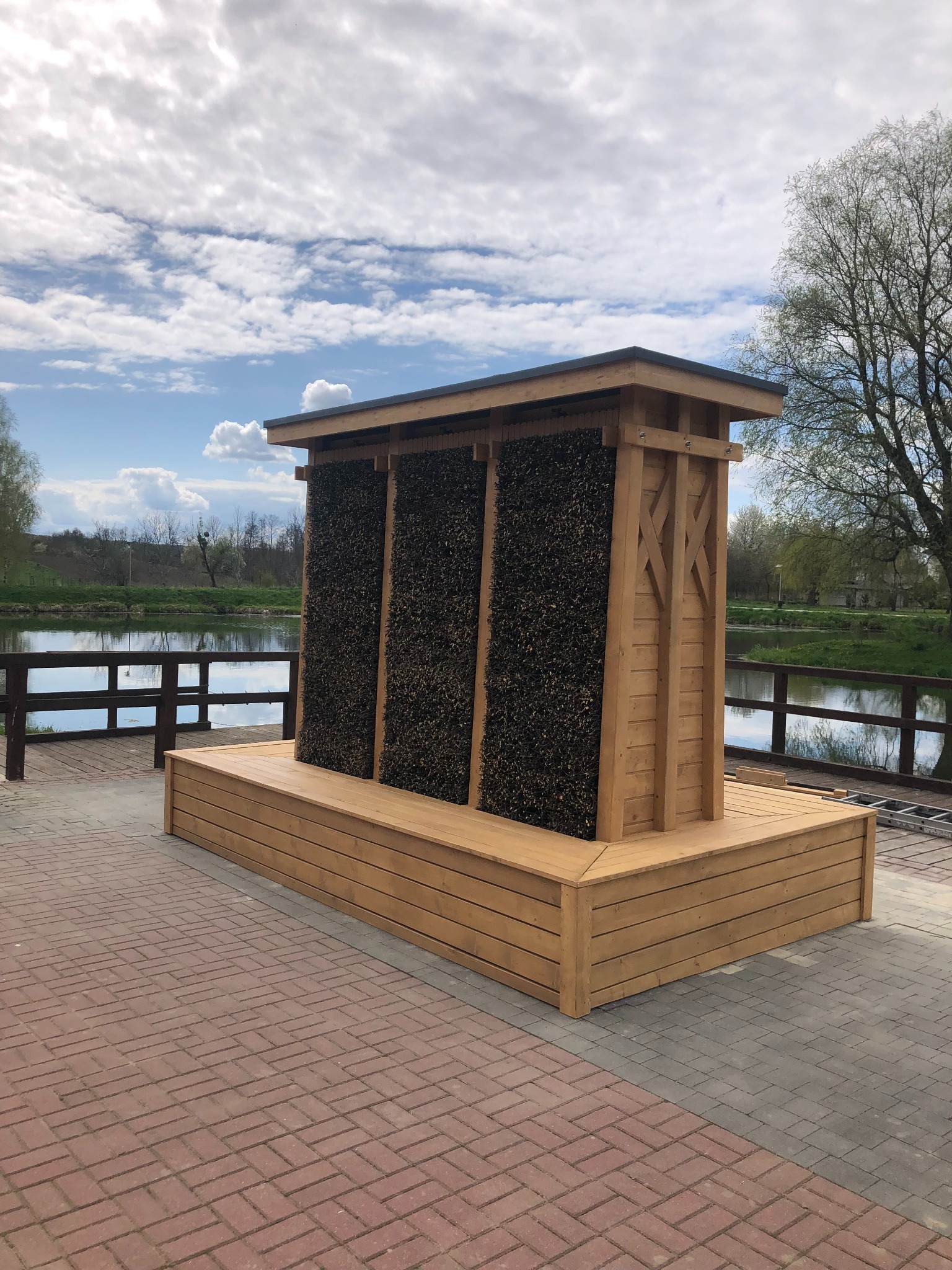 Drewniana tężnia solankowa na platformie, z widokiem na rzekę i drzewa w tle, pod błękitnym niebem z chmurami. Konstrukcja z jasnego drewna, wypełniona ciemną tarniną.