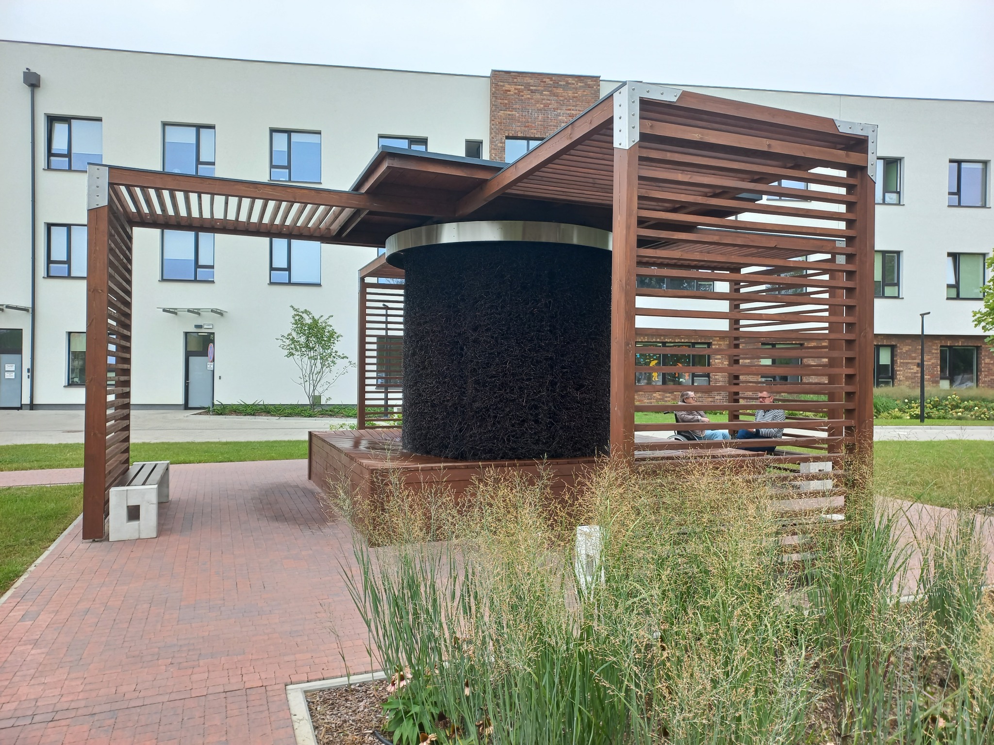 Drewniana konstrukcja solankowa z elementami pergoli na tle budynku. Widoczne osoby siedzące wewnątrz konstrukcji. Ujęcie z poziomu trawy, z roślinami na pierwszym planie.