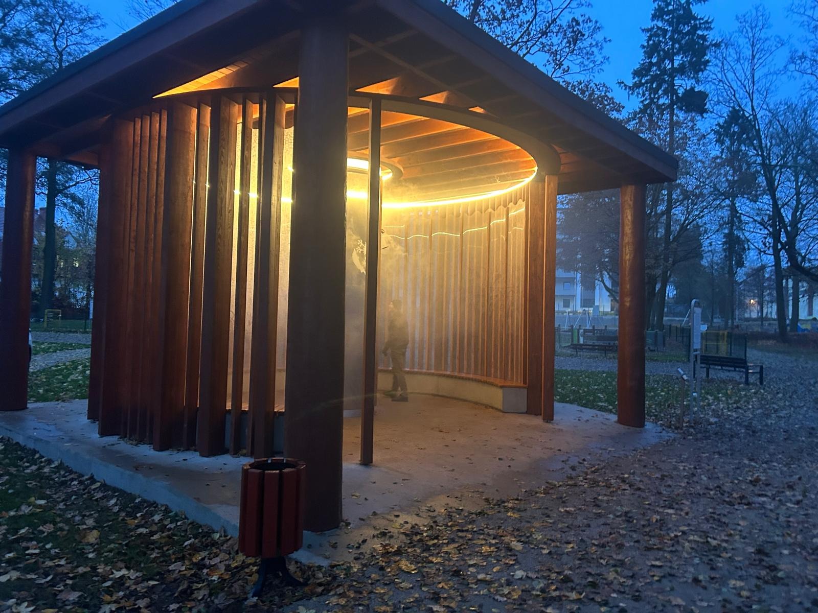 Nowoczesna altana z drewnianymi lamelami i oświetleniem LED w parku, widziana z zewnątrz, z osobą w środku. Jesienna sceneria, wieczorne światło.