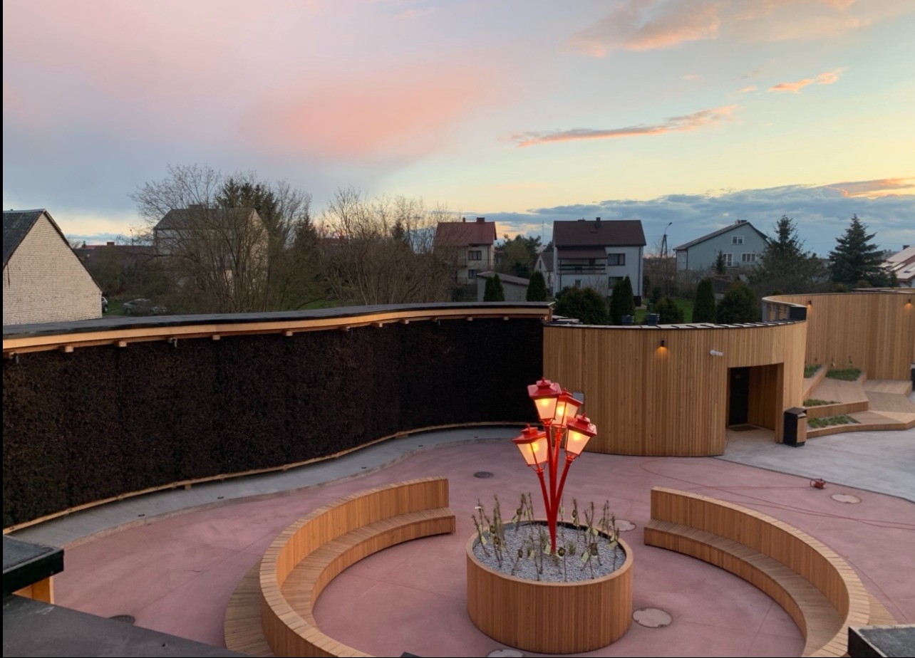 Tężnia solankowa z drewnianymi elementami i oświetleniem w kształcie lamp, otoczona zabudową z drewna na tle zabudowań mieszkalnych i nieba o zachodzie słońca.