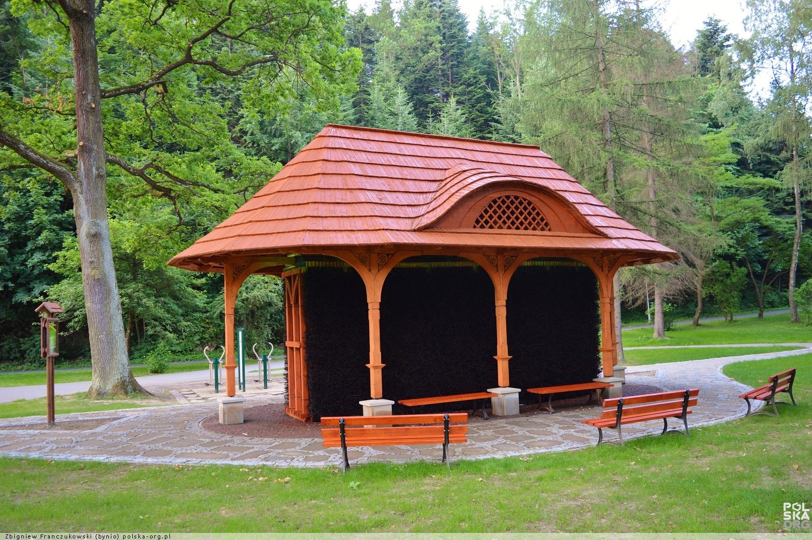 Drewniana konstrukcja tężni solankowej z charakterystycznym dachem krytym gontem, otoczona zielenią parku i ławkami. Widok na architekturę uzdrowiskową w słoneczny dzień.