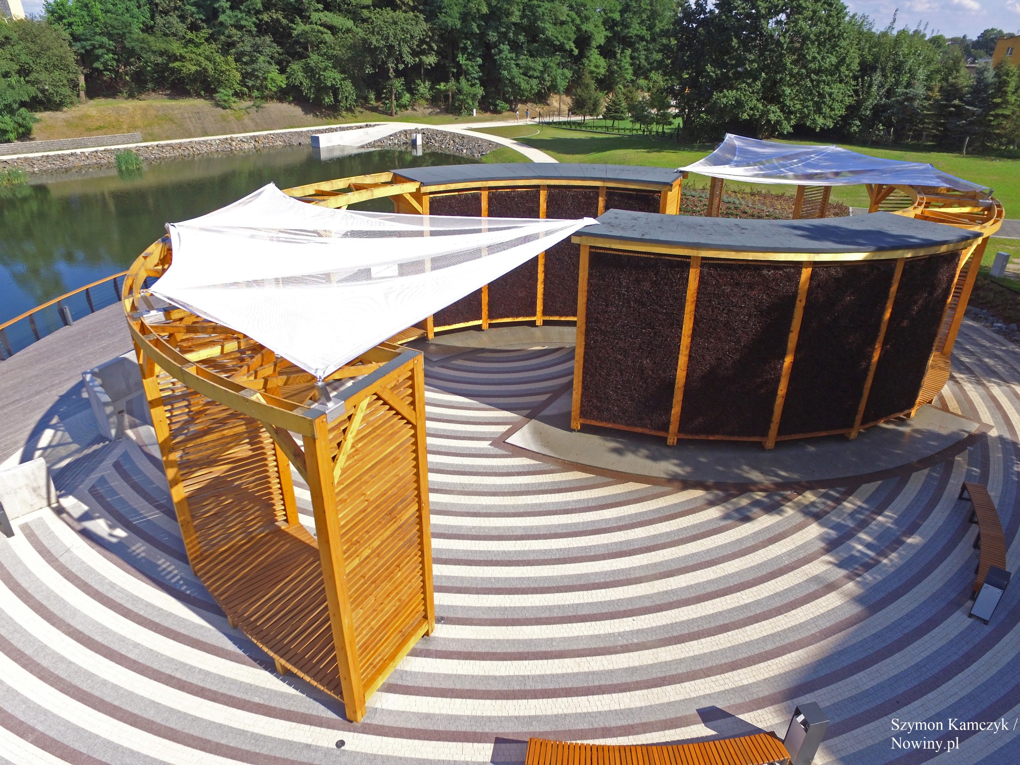 Kompleks solankowy z drewnianymi konstrukcjami i białymi żaglami, widok z góry na okrągły plac z wzorem w paski, otoczony zielenią i zbiornikiem wodnym.