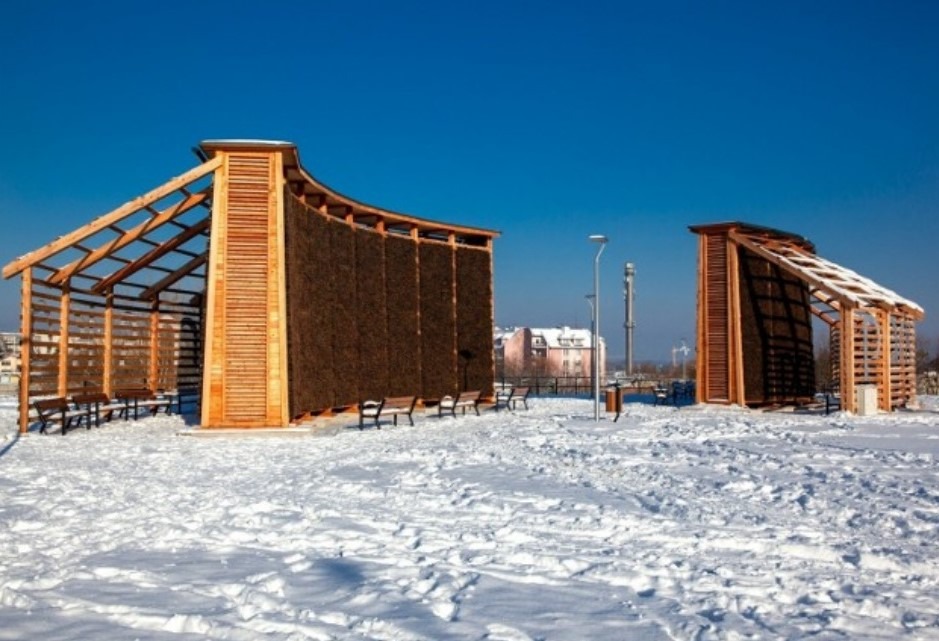 Dwie drewniane tężnie solankowe w zimowej scenerii, z widocznymi elementami konstrukcyjnymi i ścianami z tarniny. Błękitne niebo w tle.