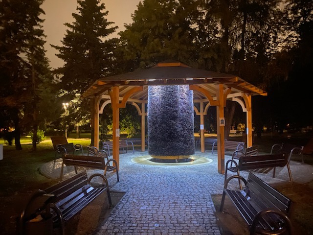 Oświetlona tężnia solankowa w drewnianej konstrukcji z dachem, otoczona ławkami i brukowaną ścieżką w nocnym parku. Unikalne ujęcie z bliska.