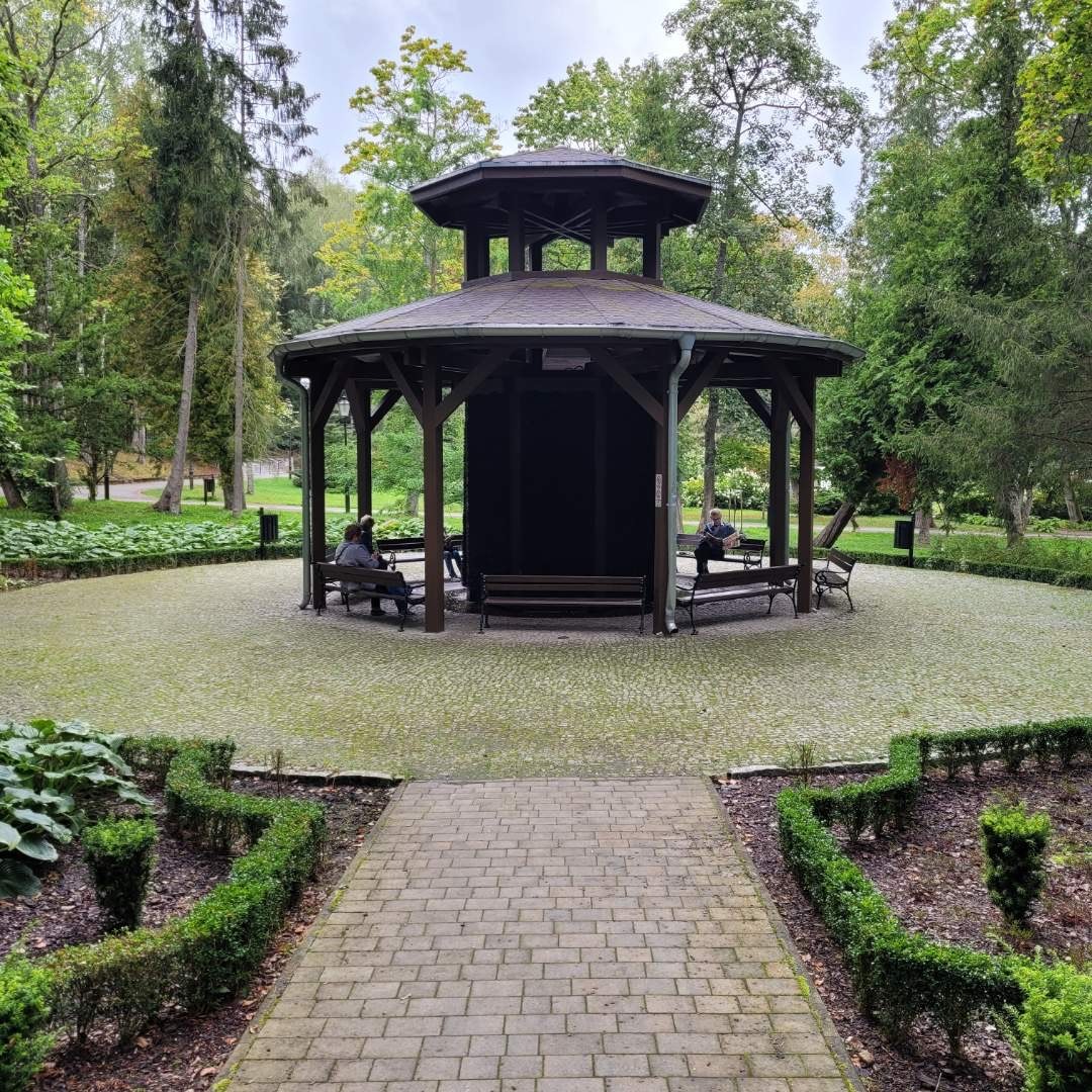 Ośmioboczna altana z ciemnego drewna w parku, z osobami siedzącymi na ławkach wewnątrz. Brukowana ścieżka prowadzi do altany, otoczonej zielenią i drzewami.